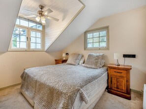 Une chambre avec un plafond en pente, un lit recouvert d’une couvre-lit à motifs, deux tables de chevet avec des lampes et une prise électrique fixée au mur.