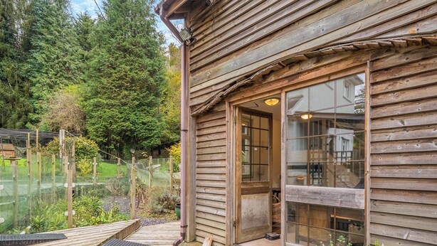 A wooden cabin with a glass door and windows, a wooden deck, and a garden area with plants and a fence. - Casa BowWow - UK48047 (Rudry, near Cardiff)
