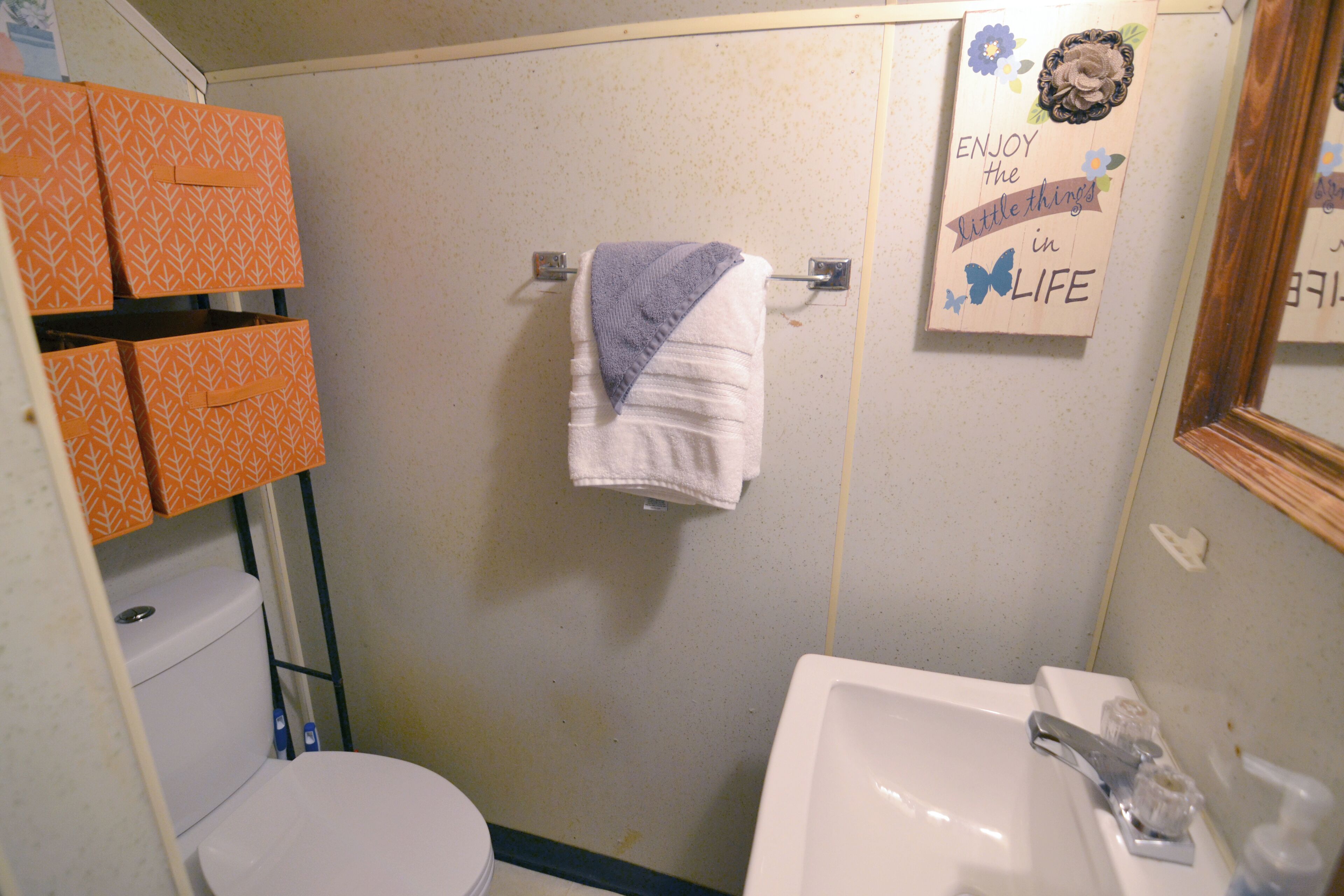 A bathroom with a toilet, sink, and towel hanging on a rack. There are orange patterned boxes on the wall and a sign that says "ENJOY the little things in LIFE".
