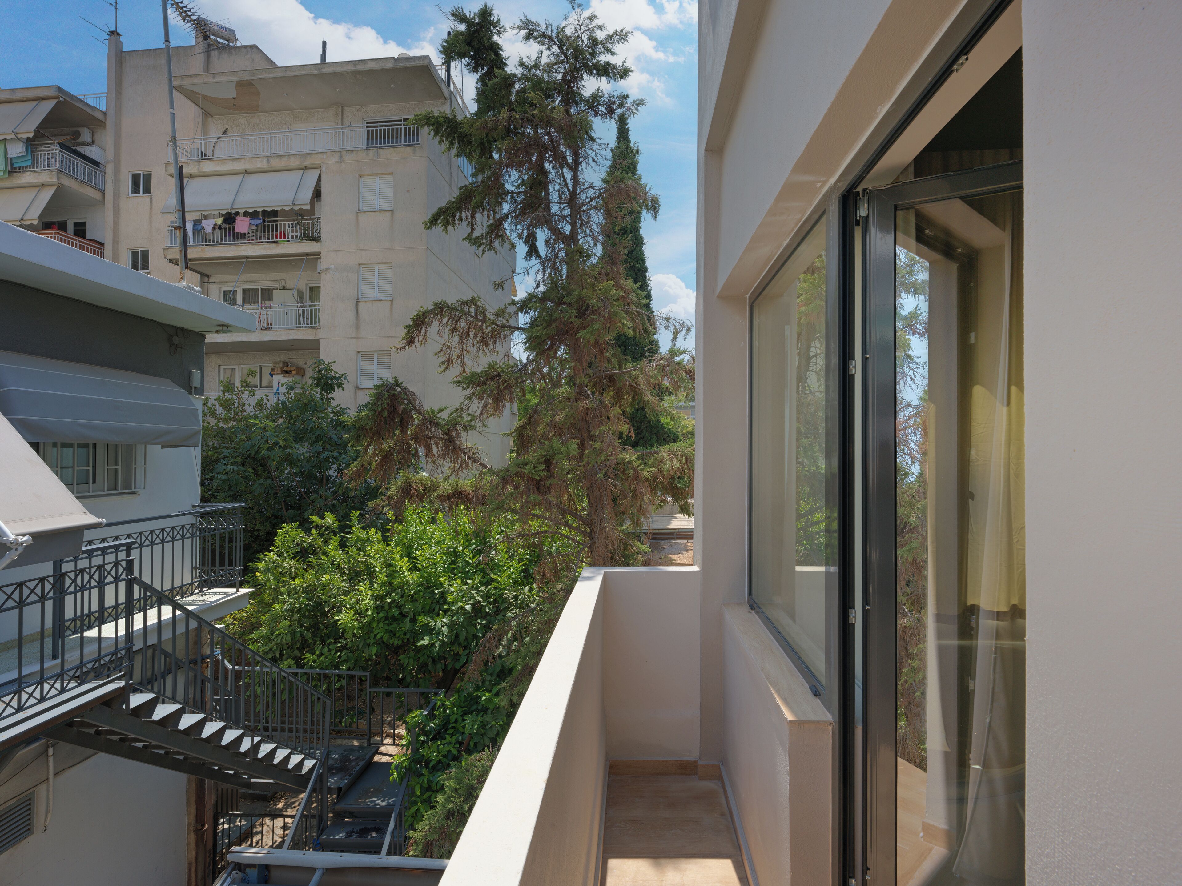 A narrow balcony with a metal railing, overlooking a courtyard with trees and adjacent buildings.