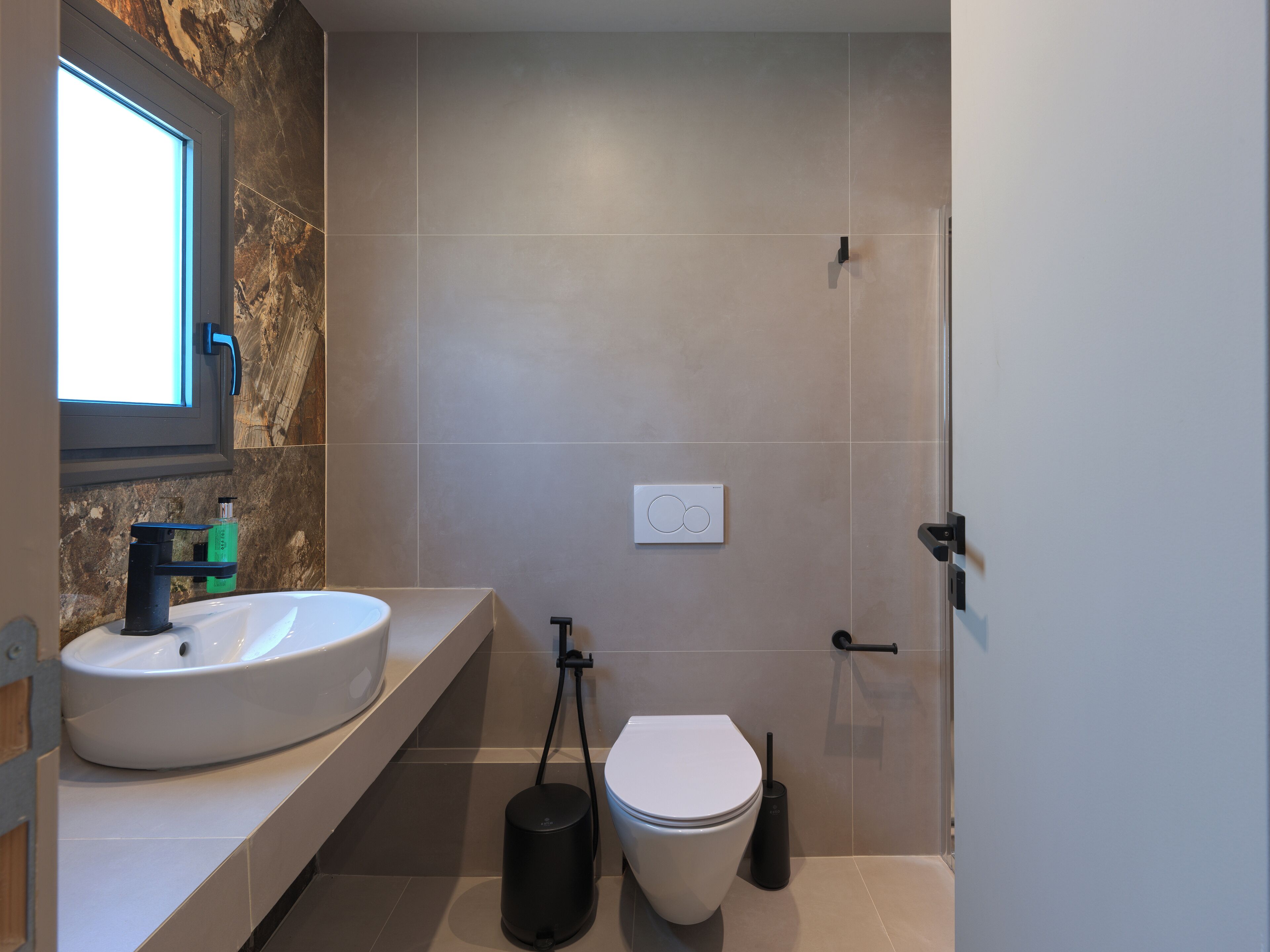 A modern bathroom with a marble wall, a white sink, a toilet, and a black handheld bidet.