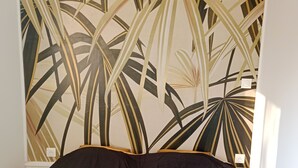 A room with a bed, a wall with a tropical leaf pattern, and two wall-mounted light fixtures. - Pounette, apartment T2 RDC, 1 bedroom facing the thermal baths, villa des Tilleuls  (Luxeuil-les-Bains)