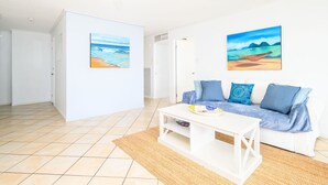 Una sala de estar con un sofá blanco, dos cojines, una mesa de centro y dos cuadros con escenas de playa.