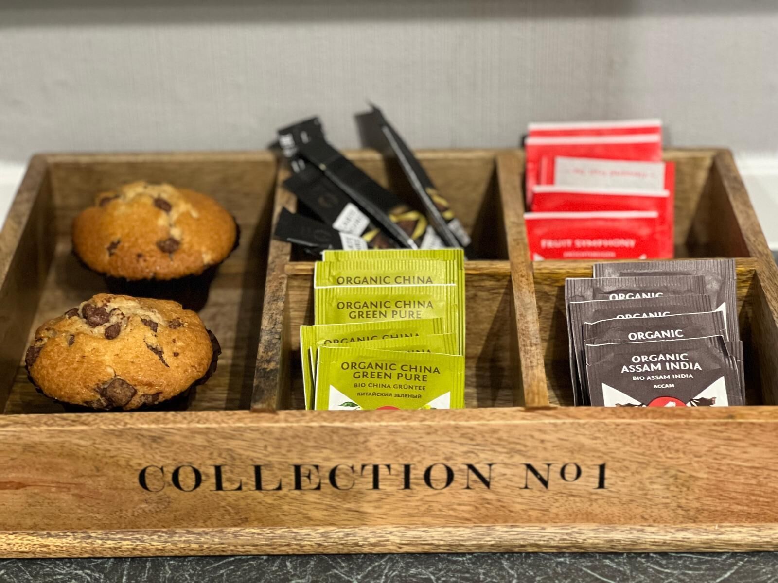 A wooden box labeled "COLLECTION No 1" containing organic tea packets, a muffin, and a pen.