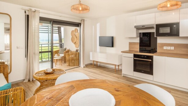 Een moderne keuken met witte kastjes, een magnetron en een fornuis. Er is een eethoek met een ronde houten tafel en witte stoelen.