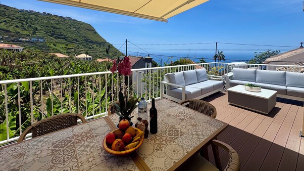Eine Terrasse mit Tisch, bedeckt mit Früchten und einer Flasche, weißen Sofas und Blick aufs Meer.