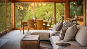 A cozy interior with a wooden dining table and chairs, a sofa with cushions, and a large window offering a view of greenery.