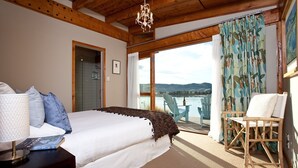 A bedroom with a large bed, a wooden ceiling, a chandelier, a view of a lake, and a chair by the window.