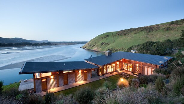 A modern house with a large deck overlooking a body of water, surrounded by hills.