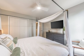 Un dormitorio con cama, ventilador de techo, televisión, cómoda y ventana con cortinas.