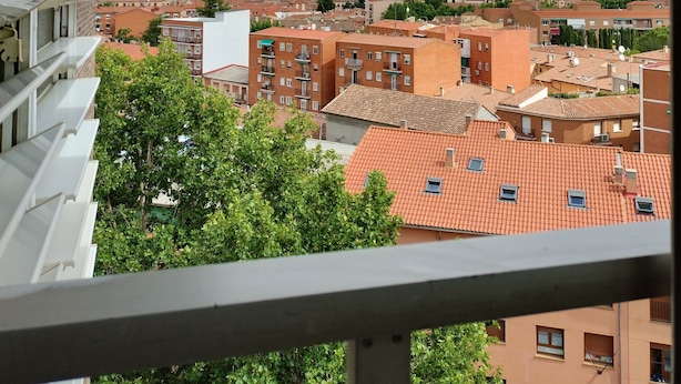 Vista desde un balcón sobre un paisaje urbano con edificios de tejados rojos, una torre destacada y cielo parcialmente nublado.