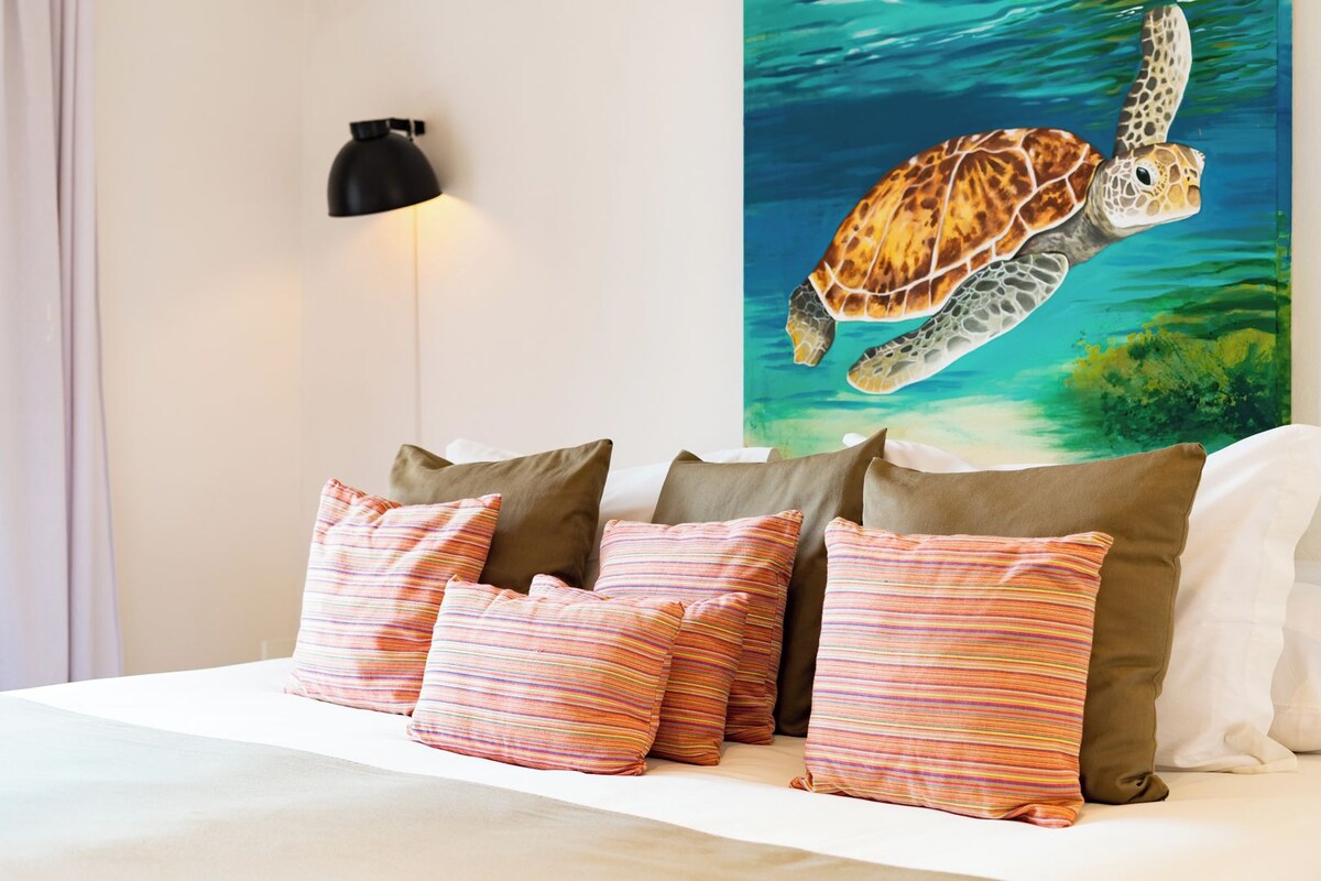 A bed with striped pillows, a wall-mounted lamp, and a painting of a sea turtle.