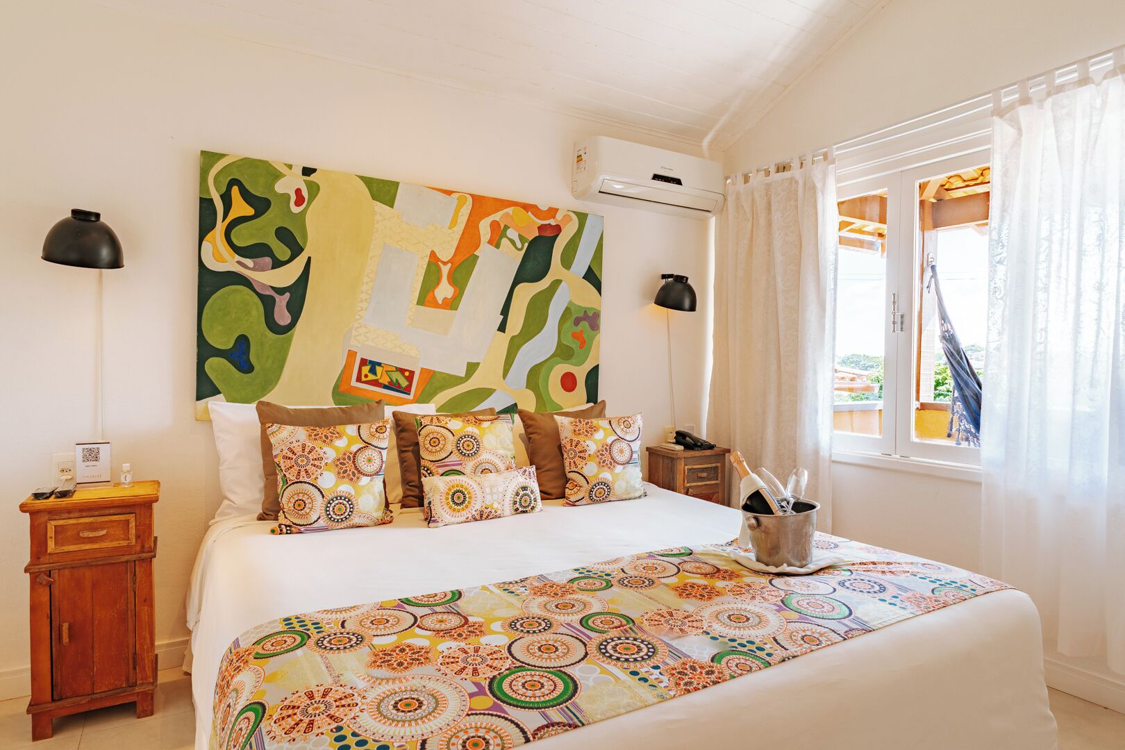 Un dormitorio con una cama grande, ropa de cama colorida, una mesita de noche y aire acondicionado instalado en la pared, además de un ventanal con cortinas.