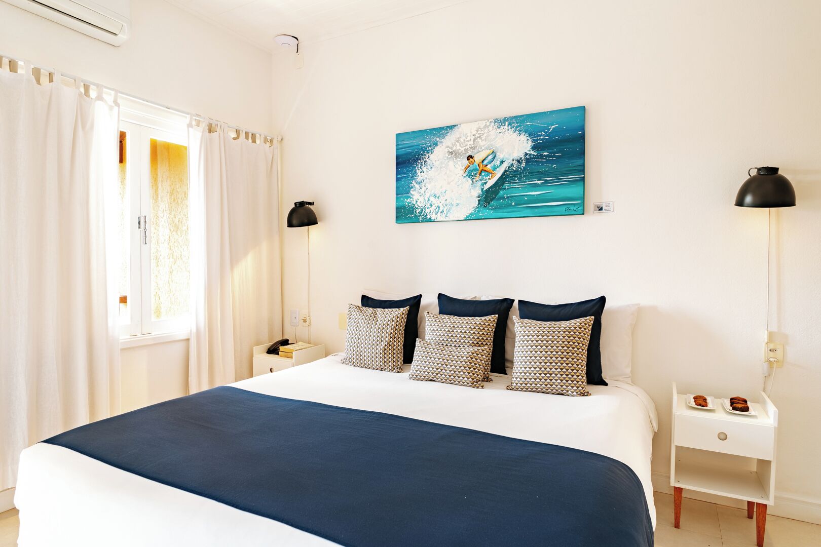 A hotel room with a large bed, a painting of a surfer on a wave, a nightstand with a lamp, and a small table with a plate of food.