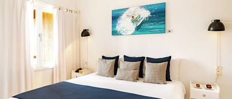A hotel room with a large bed, a painting of a surfer on a wave, a nightstand with a lamp, and a small table with a plate of food.