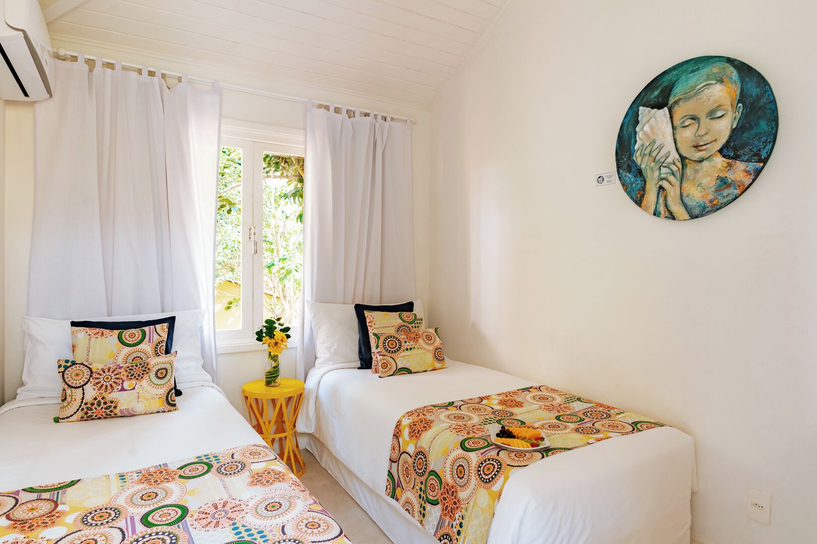 A room with two beds, each with patterned bedding and pillows, a small round table with a vase of flowers, and a painting on the wall.