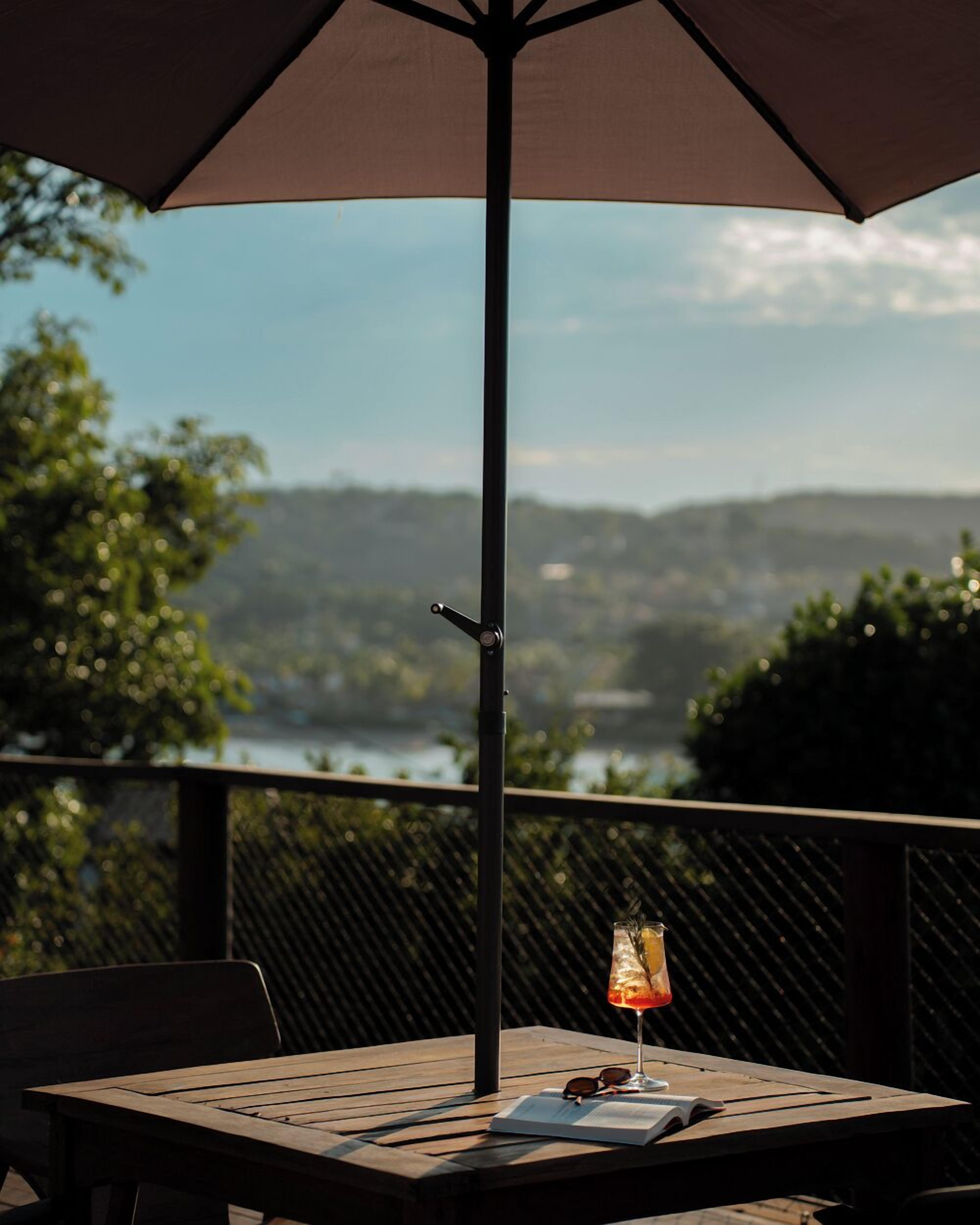 A wooden table with a drink, sunglasses, and a book under a large umbrella, overlooking a scenic view of hills and a body of water.