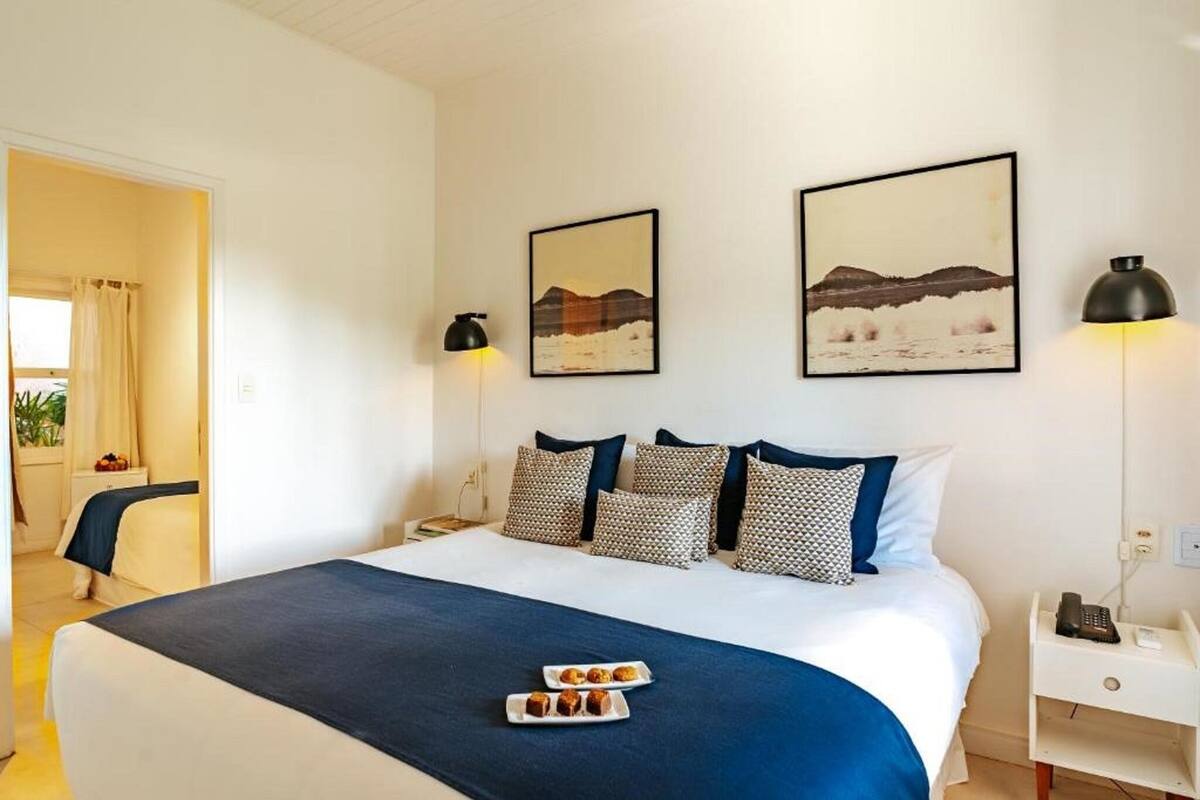 Habitación de hotel con una cama, dos lámparas de pared, dos cuadros enmarcados, una mesita de noche con un teléfono y una bandeja de pasteles sobre la cama.