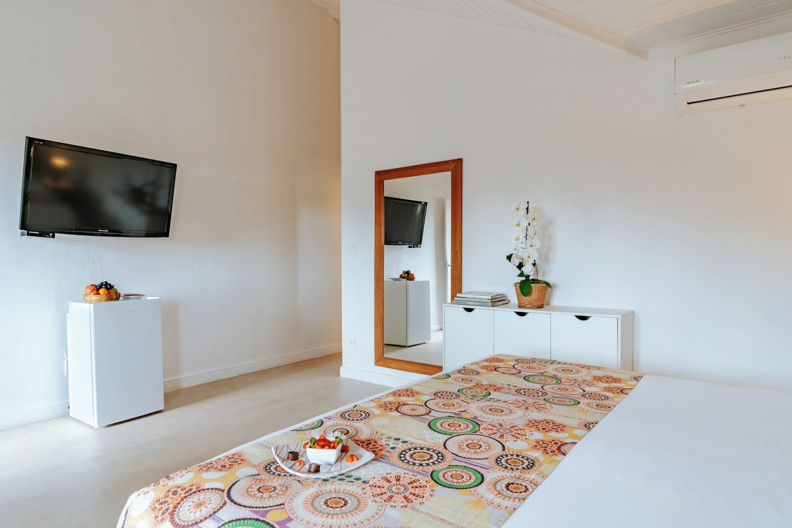 A modern bedroom with a bed, a television, a small refrigerator, a dresser, and a mirror.
