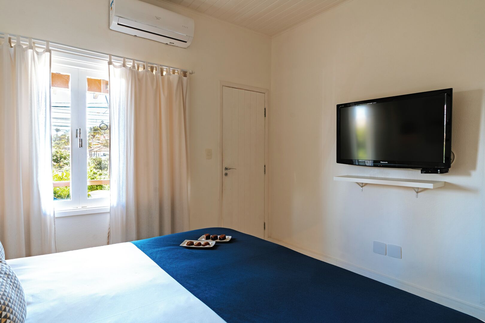 Habitación de hotel con cama, televisor en un mueble bajo, ventana con cortinas y equipo de aire acondicionado.