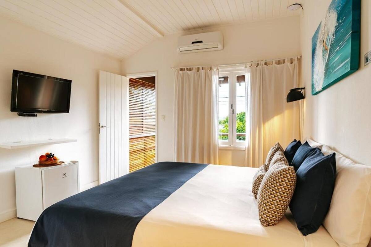 Un dormitorio con una cama, televisión, refrigerador pequeño, ventana con cortinas y un cuadro en la pared.