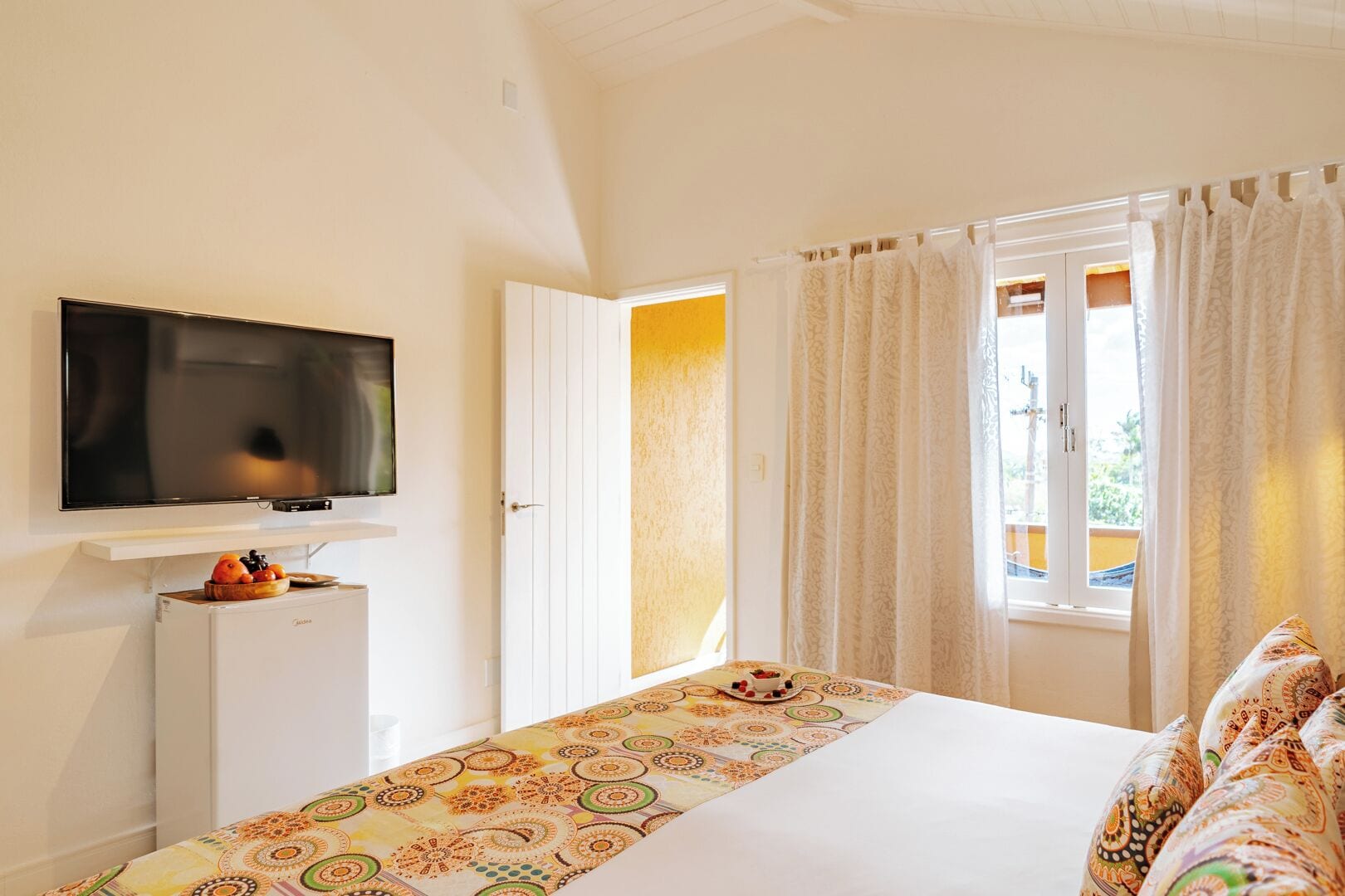 A hotel room with a bed, a TV mounted on the wall, a small fridge, and a window with curtains.