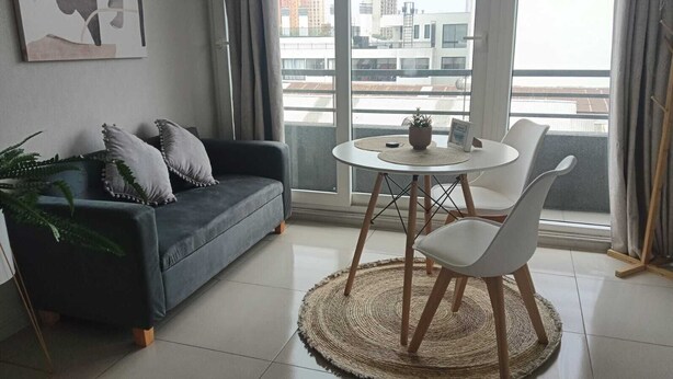 Una sala moderna con un sofá gris oscuro, sillas blancas, una mesa redonda y vistas a edificios a través de amplios ventanales.