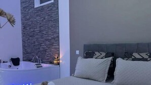 Un dormitorio con cama, almohadas, una mesita de noche, lámpara y baño con bañera y espejo.