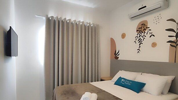 A hotel room with a bed, a flat-screen TV mounted on the wall, a window with curtains, and an air conditioning unit. - Búzios Suíte Home (Búzios)