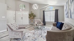 A spacious living room with a large window, a tufted armchair, a glass coffee table, and a beige sofa. - Fascinating private pool and game-room, near Disney!! (Kissimmee)