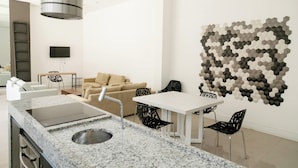 A modern kitchen with a granite countertop, a built-in stove, and a stainless steel sink. There is a dining area with a white table and black chairs. A beige sofa and a black chair are in the background. - Flat Vip 5 Estrelas (Cotia)