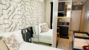 A hotel room with a modern design, featuring a geometric-patterned wall, a white bed with patterned pillows, a small desk, and a kitchenette in the background. - Flat Vip 5 Estrelas (Cotia)