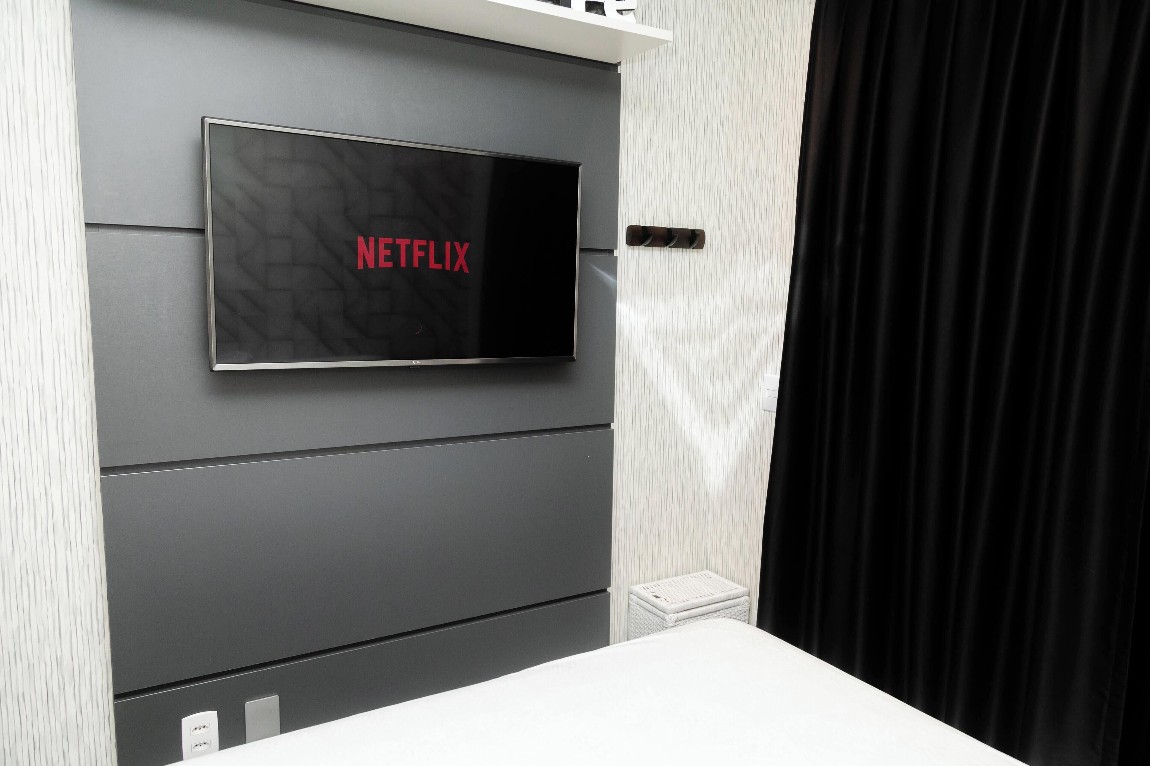 Een moderne kamer met een flatscreen televisie aan een grijze muur, een witte nachtkastje en een zwart gordijn.