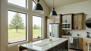 A modern kitchen with a large island, wooden cabinets, stainless steel appliances, and a view of a green landscape through large windows. - NEW charming 3 bedroom cottage nestled on the scenic Red Lodge Golf Course  (Red Lodge)