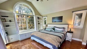 A bedroom with a large window, built-in shelving, a bed with a striped bedspread, and a nightstand with a lamp. - Lakefront home with hot tub, beach, skiing nearby in tranquil Barnstead (Barnstead)
