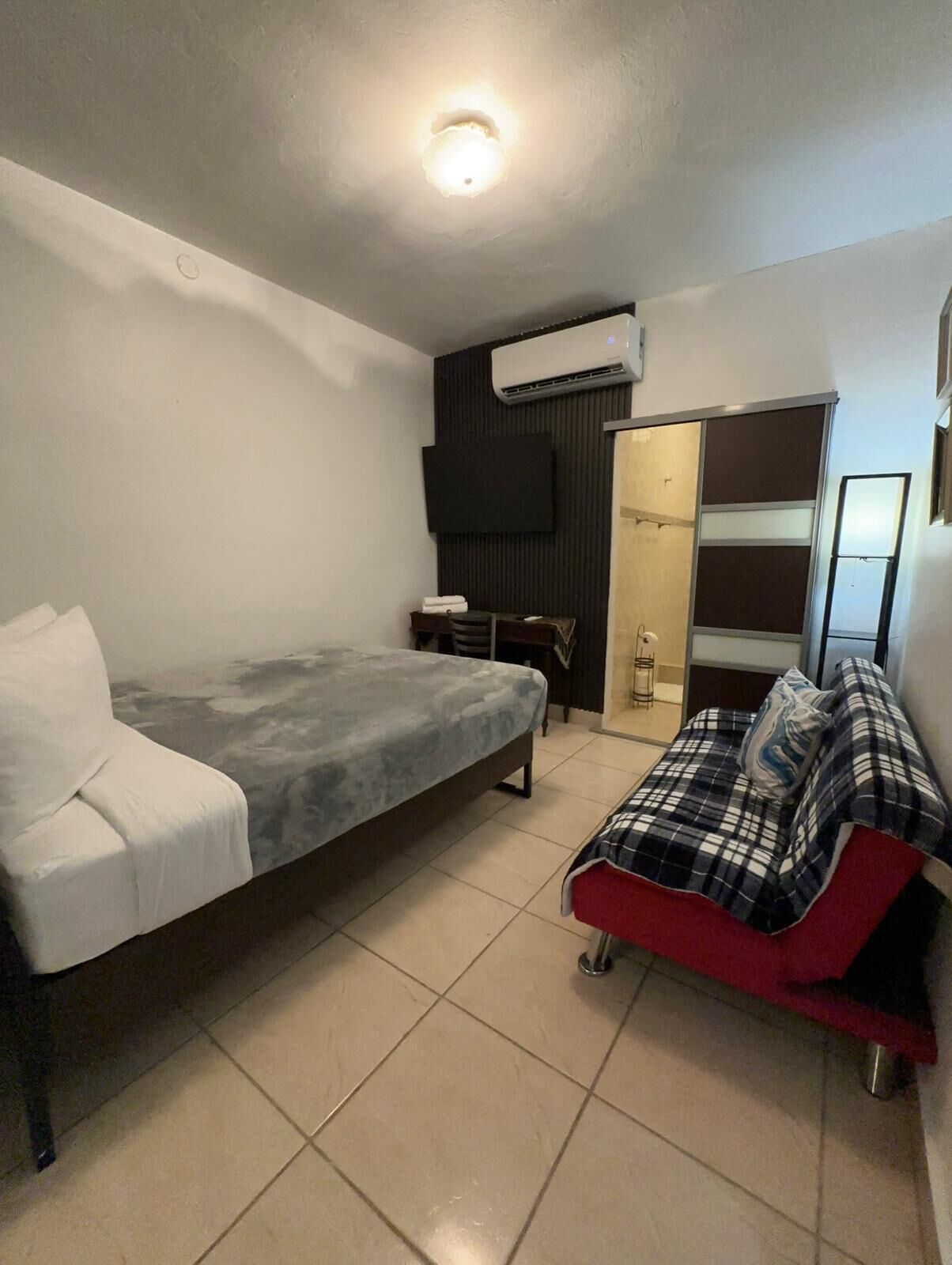 A small, modern bedroom with a bed, a sofa, a TV, and an air conditioning unit.