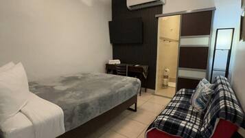 A small, modern bedroom with a bed, a sofa, a TV, and an air conditioning unit.