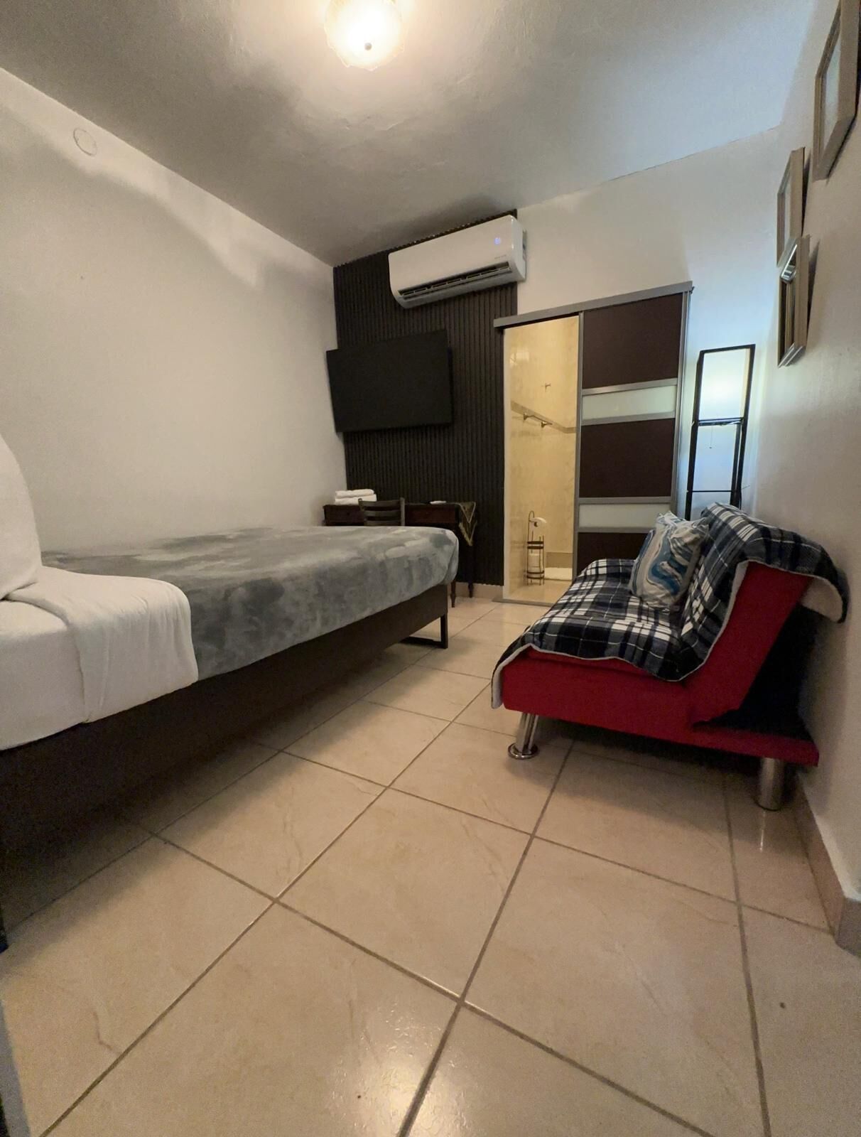 A bedroom with a bed, a red sofa, a TV, and an air conditioning unit.