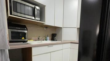 Een kleine keuken met witte kastjes, een zwarte koelkast, een magnetron, een koffiezetapparaat en een prullenbak.