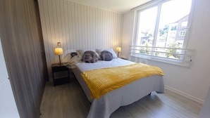 Quarto com cama, duas luminárias de cabeceira, janela e vista para prédios.