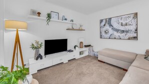 Una sala de estar con un televisor de pantalla plana, un sofá color beige, una lámpara de pie y un estante en la pared con objetos decorativos.