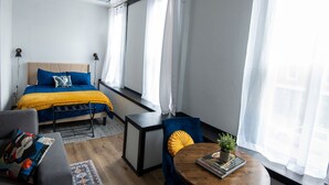 A hotel room with a bed, a desk, a chair, a small table with a plant, and a window with curtains. - Cozy studio in enchanting downtown Warrensburg with full kitchen, AC, WiFi (Warrensburg)