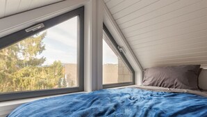 Ein Schlafzimmer mit großem Fenster, einem Bett mit blauer Decke und einer schrägen Decke.