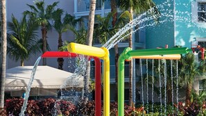 Una fuente con múltiples caños y chorros de agua de colores, rodeada de palmeras y un edificio con balcones.