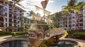 Ein Wasserspiel mit einer bootsförmigen Struktur in einem Pool, umgeben von Palmen und Hotelgebäuden.