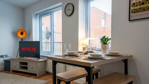 Una sala moderna con una mesa de comedor lista para cuatro, un televisor mostrando 'NETFLIX' y una planta en el alféizar de la ventana.