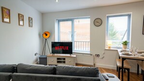 Una sala moderna con un sofá gris, un televisor sobre un mueble bajo, una mesa de comedor lista para cuatro personas y una ventana con persianas.