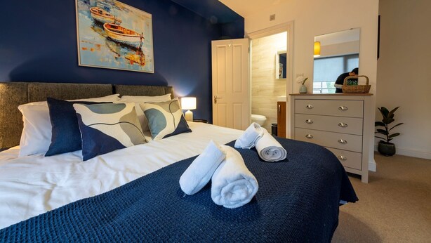 Una cama bien hecha con ropa de cama blanca y azul, un cuadro de barcos en la pared, un aparador con cajones y una planta en maceta.