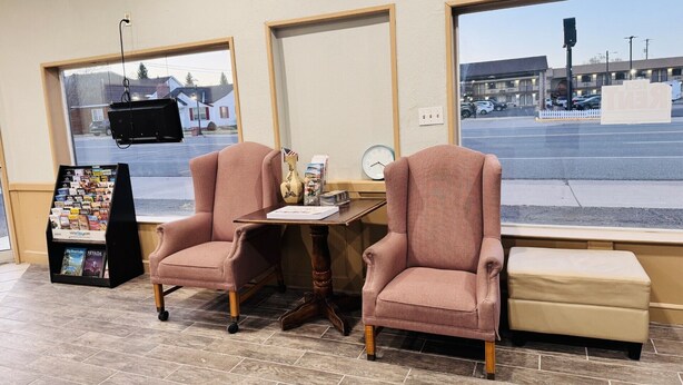 A seating area with two chairs and a small table, a clock, and a window with a view of a street and buildings. - Bryce Park Inn (Panguitch)