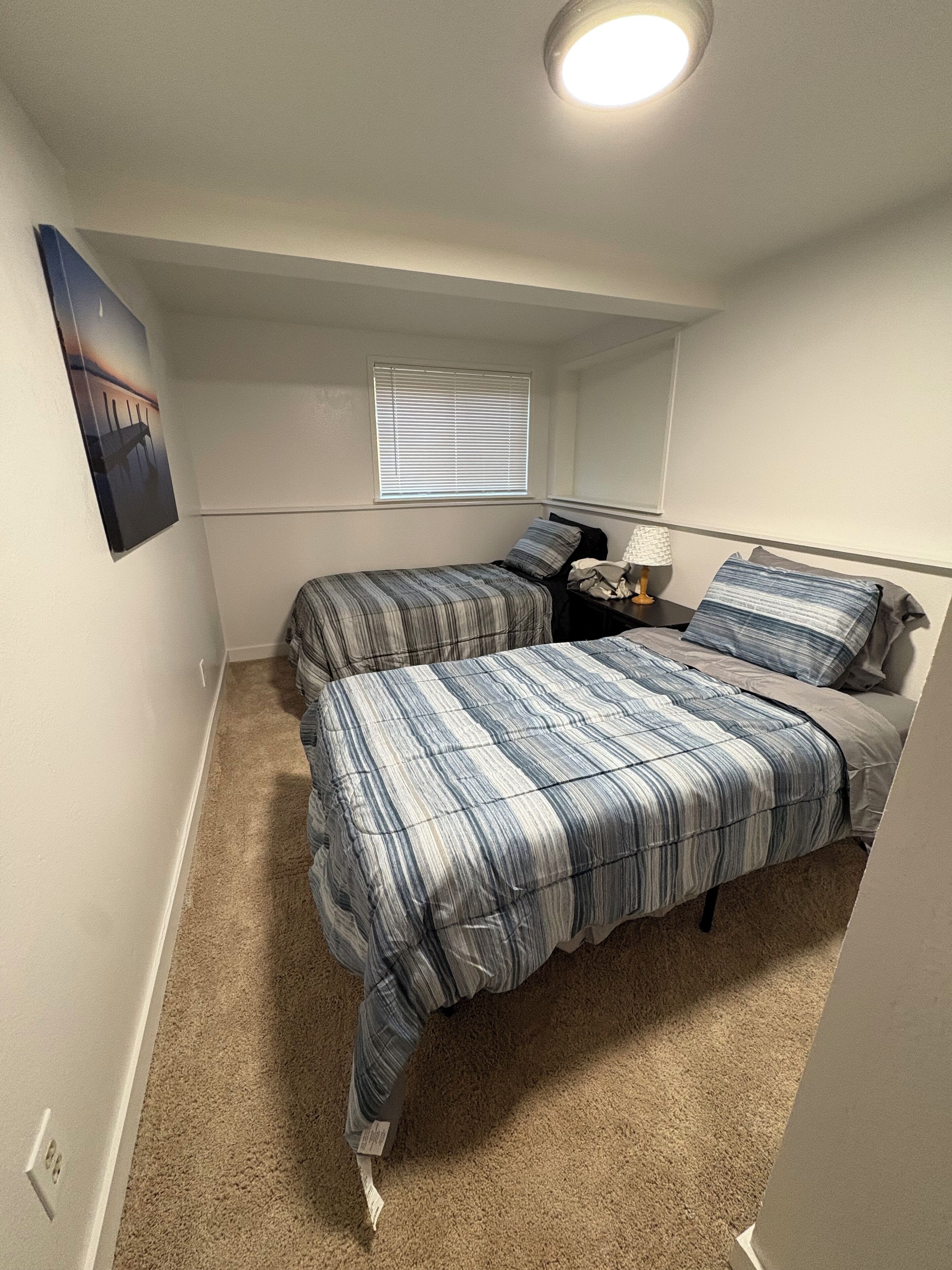 Une chambre avec deux lits, une fenêtre avec des stores, une table de chevet avec une lampe et un tableau accroché au mur.